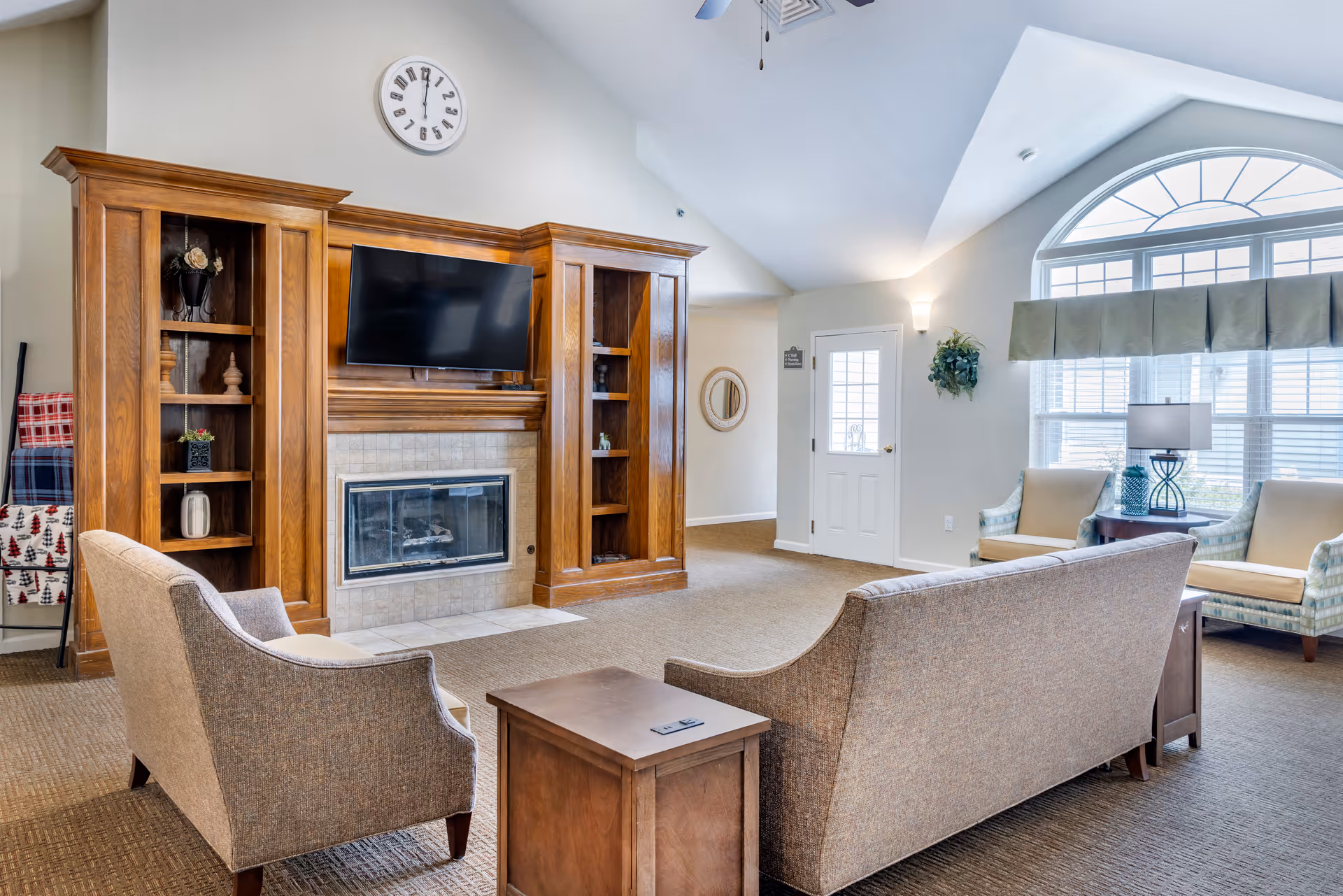 A cozy living room area with a beige sofa and armchair facing a wooden entertainment center with a mounted flat-screen TV above a fireplace. The room has a large arched window with blinds and valance, two additional armchairs with a side table and lamp, a wall clock above the fireplace, and a door leading outside. The walls are light-colored and the floor is carpeted.