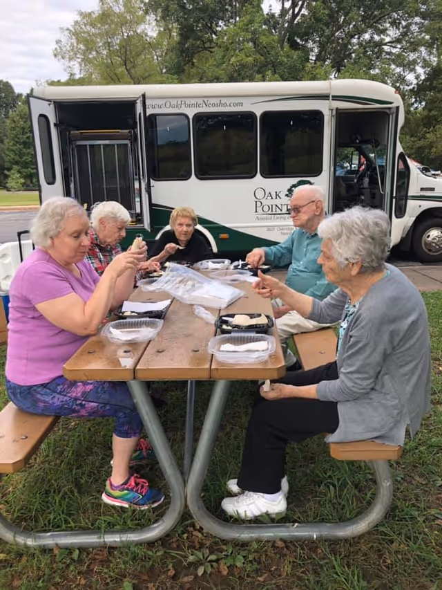 Five elderly people sitting at a wooden picnic table outdoors, eating food from takeout containers. Behind them is a white and green Oak Pointe of Neosho shuttle bus with its door open. The setting appears to be a park or grassy area with trees in the background.