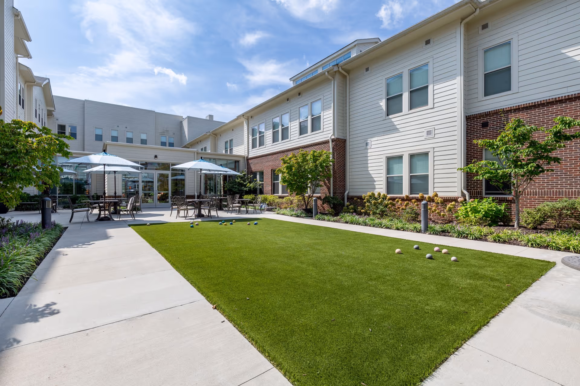 Outdoor courtyard area at Arbor Terrace Prince William Commons featuring a bocce ball court with balls scattered on the green turf, surrounded by concrete walkways. There are several tables with chairs and blue umbrellas near the building entrance. The building has white siding with brick accents and multiple windows. Trees and shrubs line the edges of the courtyard under a partly cloudy sky.