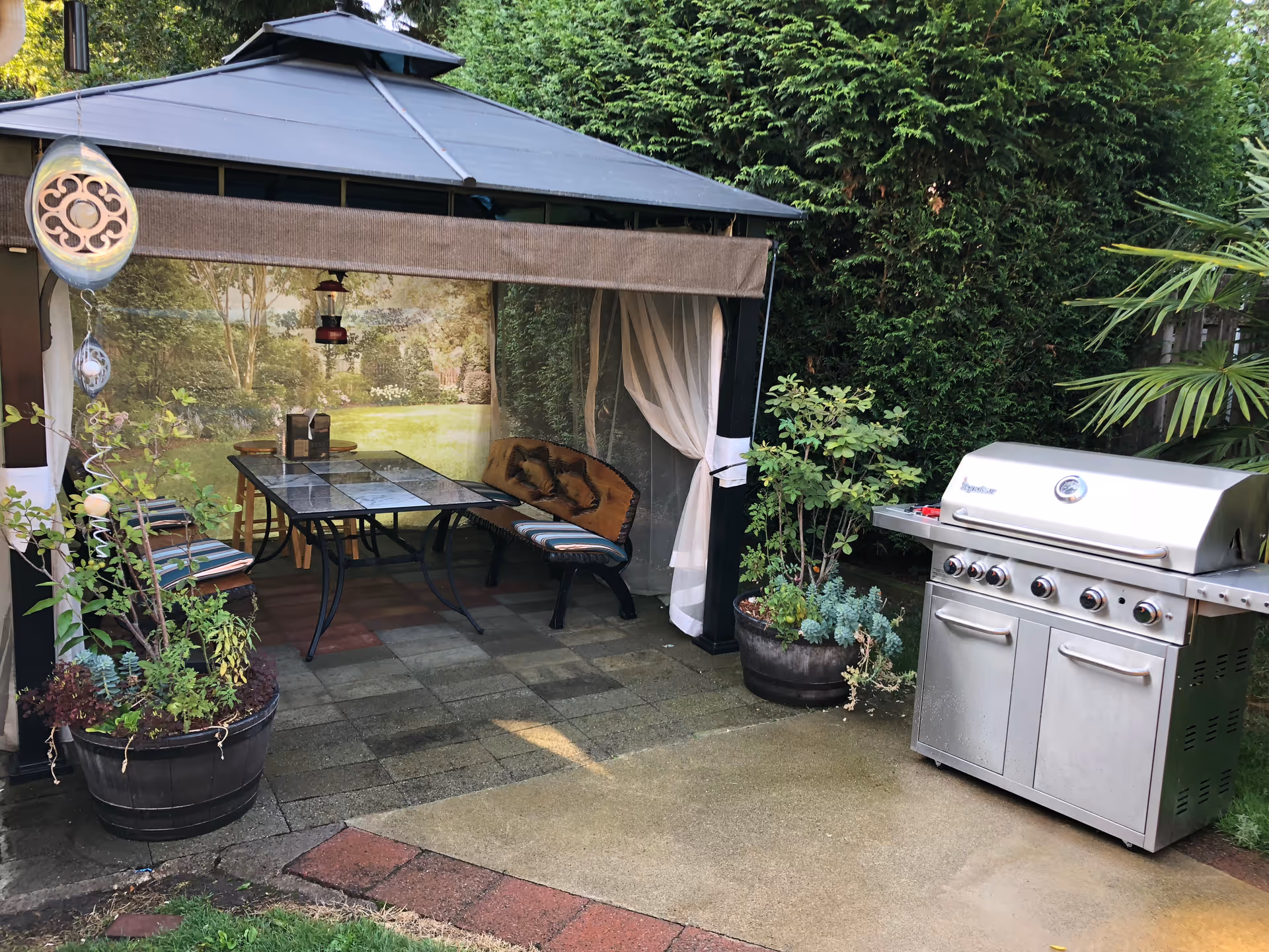 Outdoor patio area with a gazebo featuring a glass-top table and cushioned bench seating, surrounded by potted plants. To the right, there is a large stainless steel barbecue grill. The background shows tall green hedges and trees.