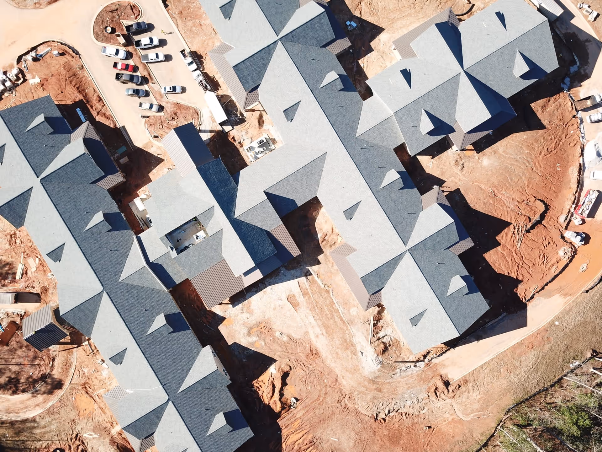 Aerial view of a senior living facility under construction showing multiple buildings with grey roofs, surrounding dirt and construction vehicles parked nearby.