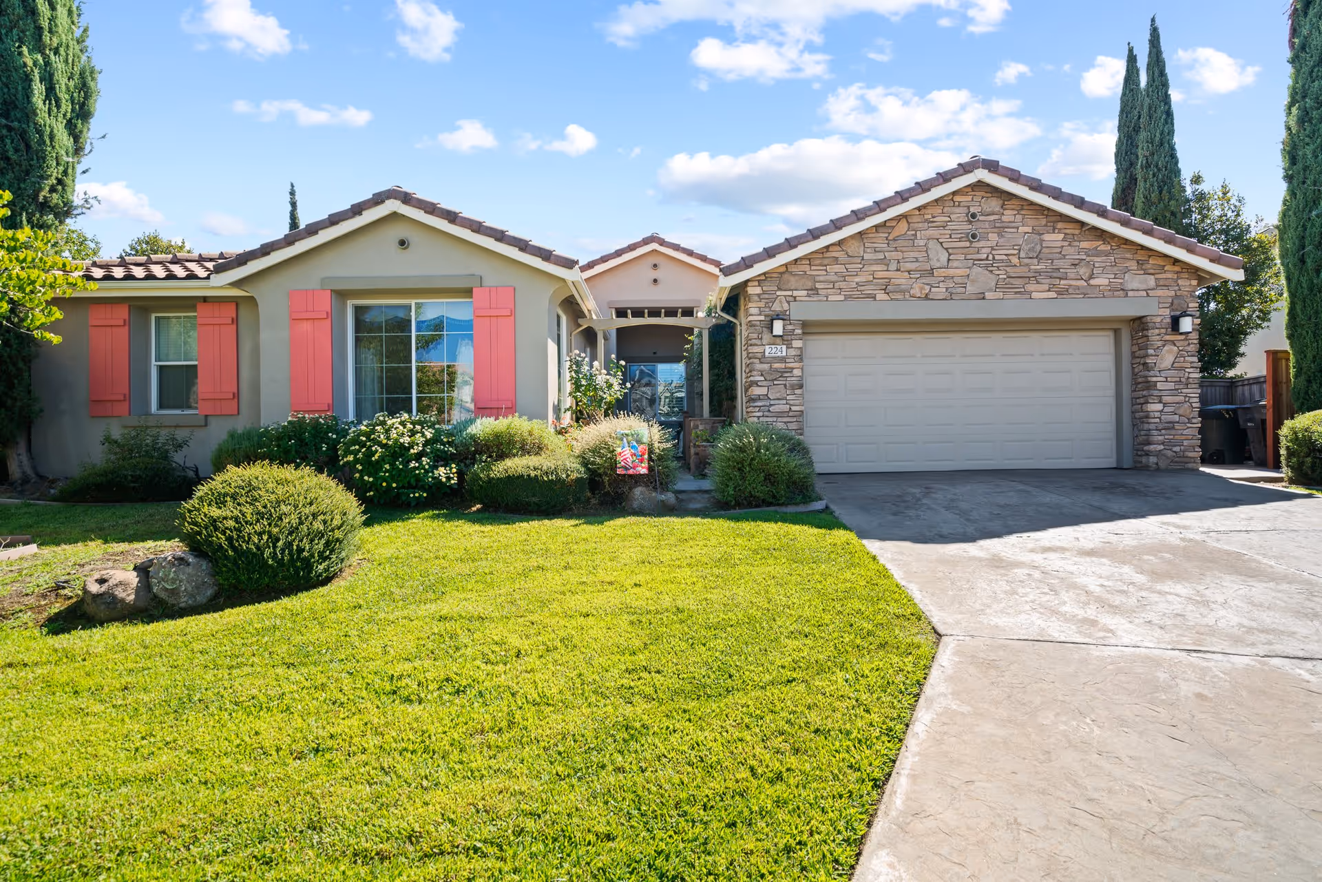 Front exterior view of a single-story house with a well-maintained green lawn, stone and stucco facade, red window shutters, and a two-car garage under a blue sky with some clouds.