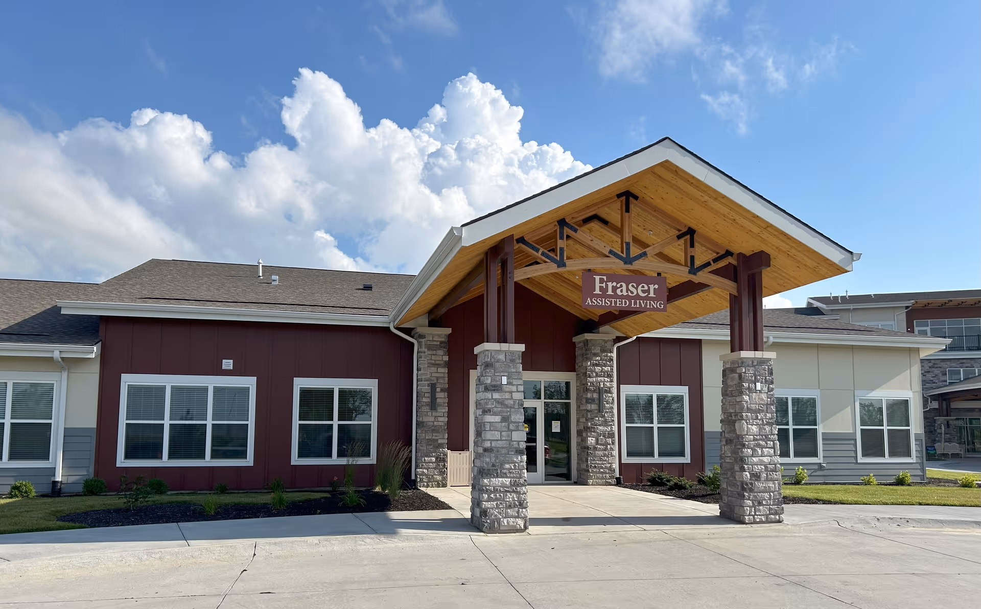 Exterior front view of Fraser Assisted Living building with a covered entrance supported by stone pillars, under a partly cloudy blue sky.