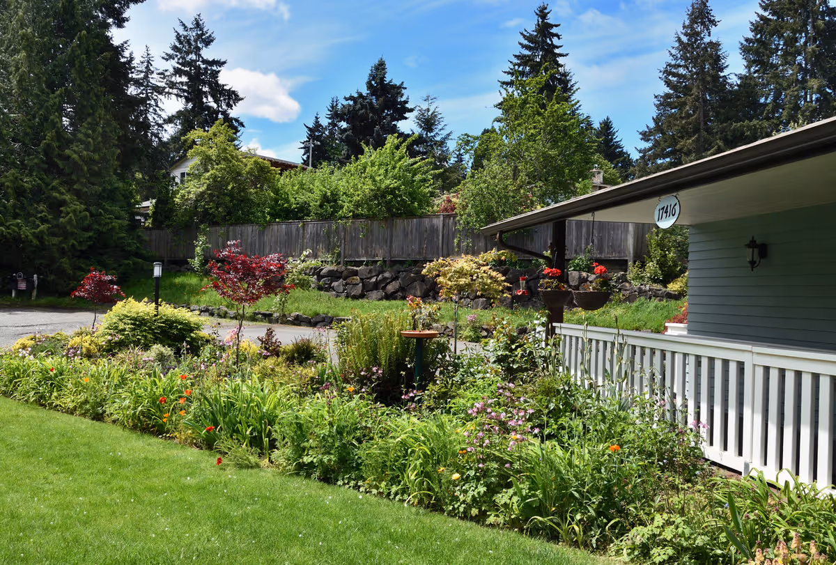 A well-maintained garden area with various green plants, flowers, and small trees in front of a building with a white railing and a sign displaying the number 17416. Tall trees and a wooden fence are visible in the background under a blue sky with some clouds.