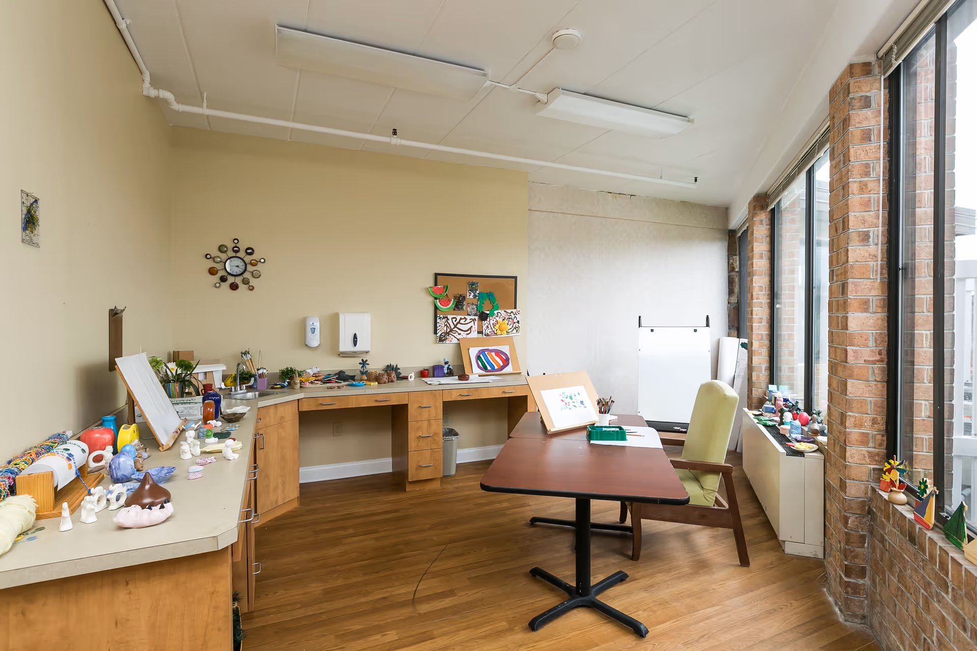 A bright activity room with large windows along one wall, wooden floors, and beige walls. The room features a long counter with various craft supplies and small art pieces displayed. There is a table with an art easel and a green chair facing the windows. The room has a clock on the wall and a bulletin board with colorful artwork pinned to it.