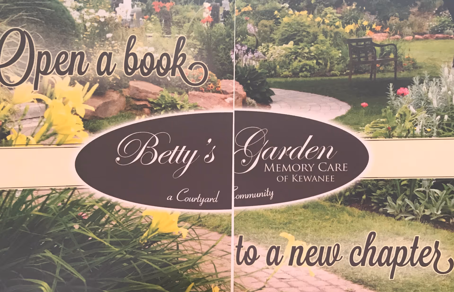A landscaped garden with a stone pathway winding through green grass, colorful flowers, and plants. There is a black metal bench in the background. Overlaid text reads 'Open a book to a new chapter' and 'Betty's Garden Memory Care of Kewanee, a Courtyard Community.'