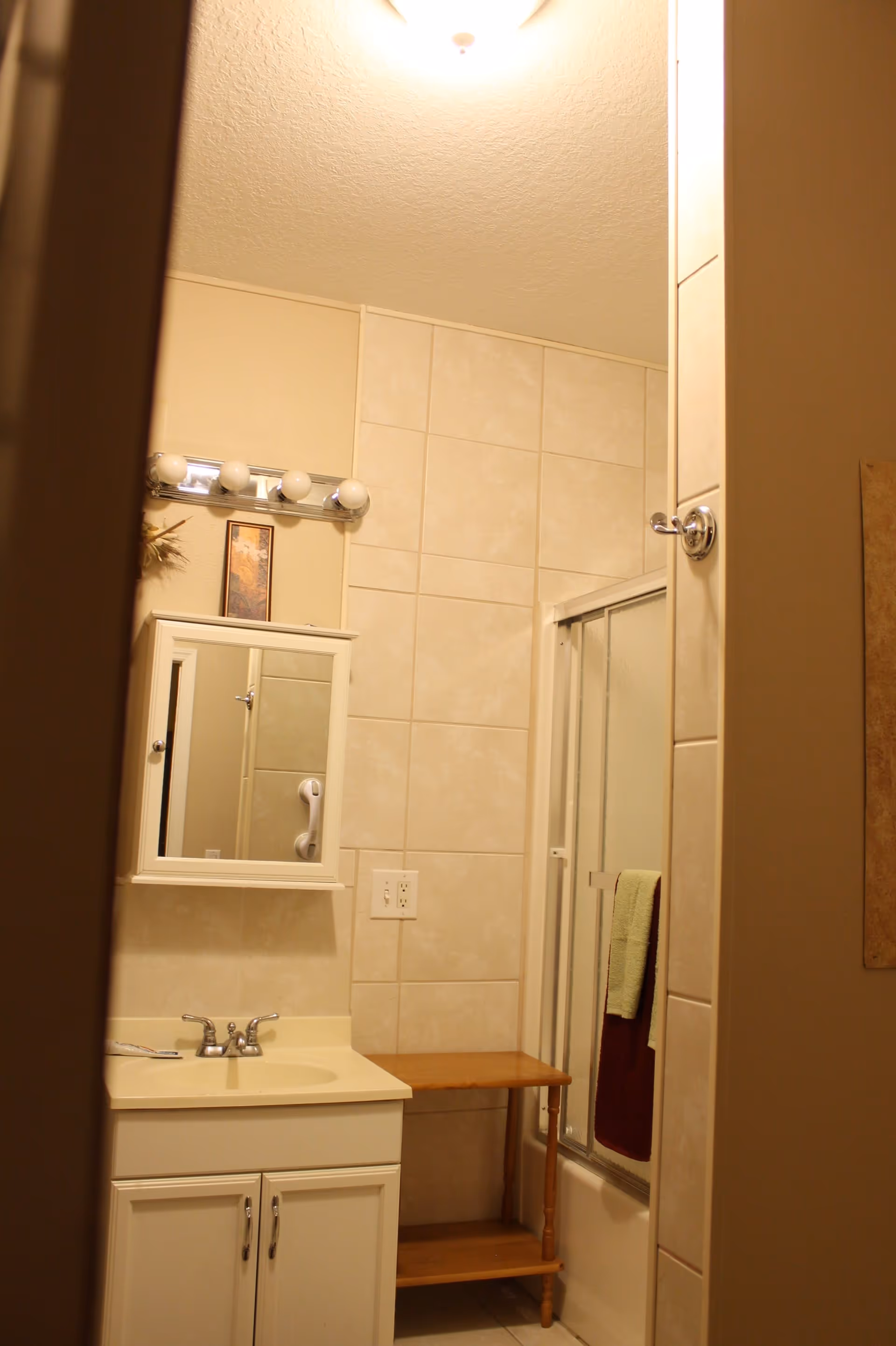 A small bathroom with beige tiled walls, a white sink vanity with a mirror cabinet above it, a wooden side table, and a shower with sliding glass doors. Two towels, one green and one burgundy, hang on the shower door. A light fixture with four bulbs is mounted above the mirror.