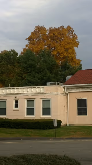Exterior view of a single-story building with beige walls and a red roof section, surrounded by green bushes and trees with autumn-colored leaves under a cloudy sky.