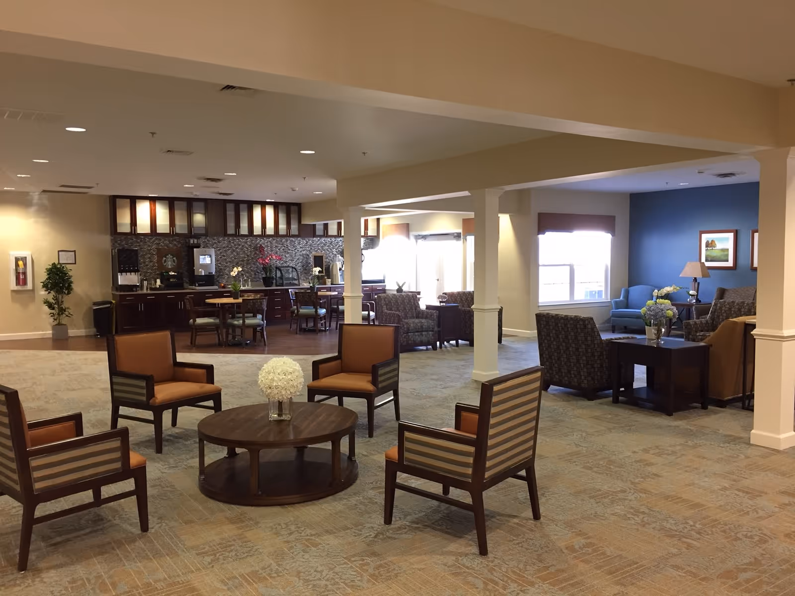 A spacious senior living facility common area with multiple seating arrangements including armchairs around a round wooden table with a flower vase. In the background, there is a kitchenette area with cabinets, a coffee machine, and a counter with chairs. The room has beige walls, carpeted floors, and large windows allowing natural light.