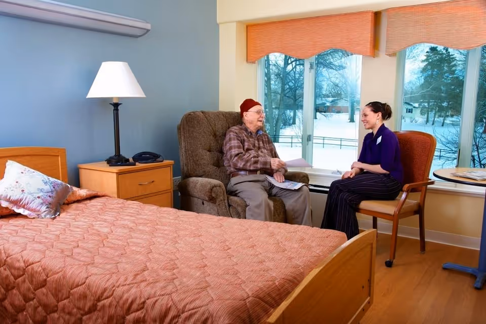An elderly man wearing a red hat and plaid shirt sits in a brown armchair next to a window with a snowy outdoor view. A woman in a purple top and striped pants sits on a chair facing him, smiling and engaging in conversation. The room has a bed with a peach-colored quilt, a wooden nightstand with a lamp and a phone, and light blue walls.