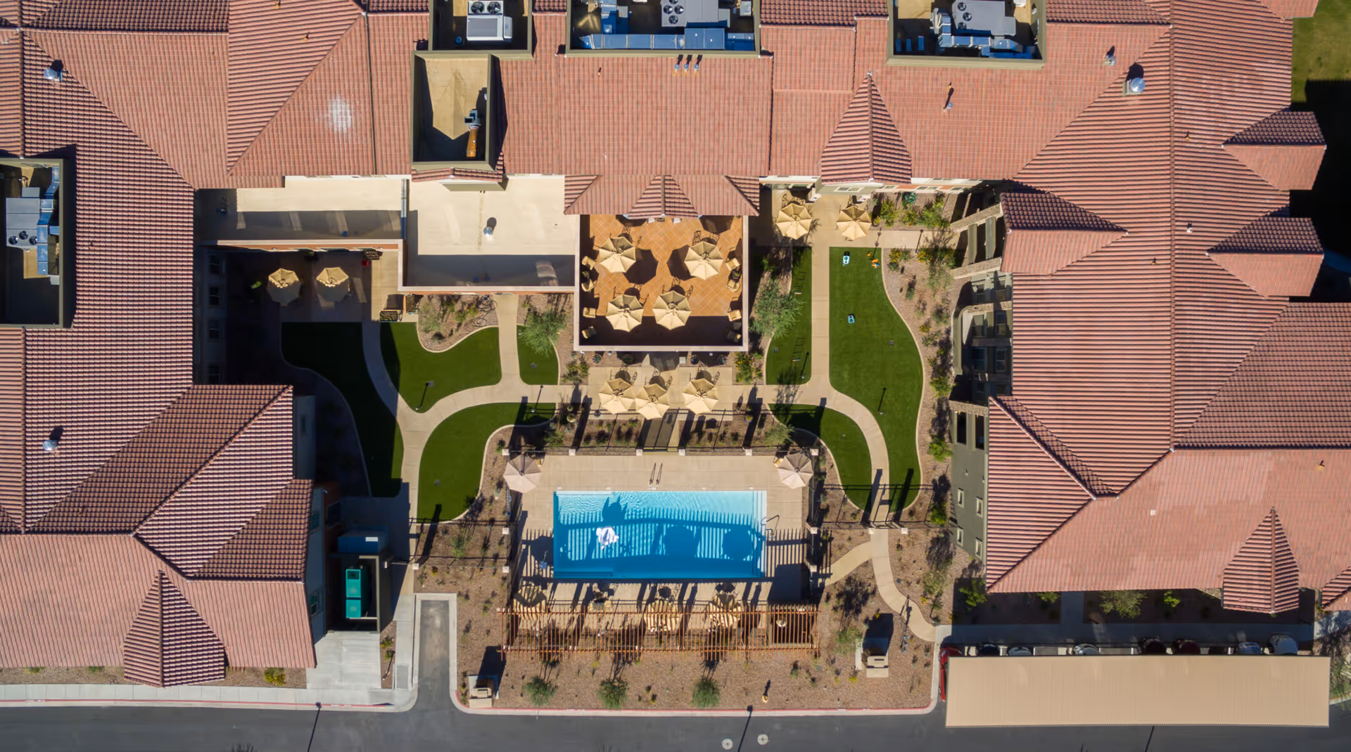 Aerial view of The Enclave at Anthem Senior Living showing a central outdoor courtyard with a swimming pool, multiple patio tables with umbrellas, green lawns, and surrounding buildings with red tile roofs.