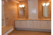 A spacious bathroom featuring a long vanity with two sinks, large mirrors above each sink, white cabinetry with multiple drawers and cupboards, warm beige tiled walls, and two wall-mounted light fixtures above the mirrors.
