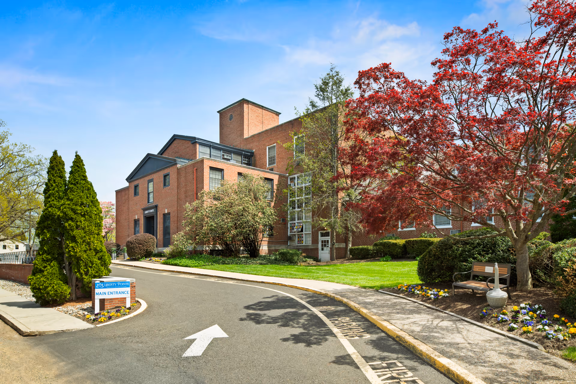 Exterior view of Liberty Pointe Rehabilitation and Healthcare Center, a multi-story brick building surrounded by green trees and landscaped gardens with colorful flowers. A paved driveway with a white arrow leads to the main entrance, and a sign indicating the main entrance is visible near the driveway.