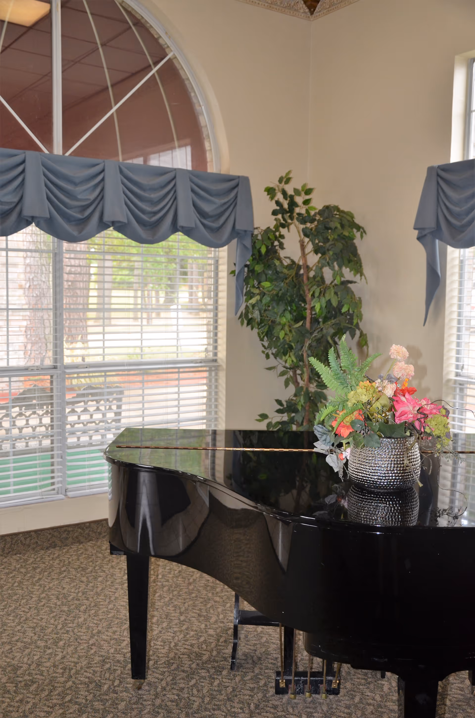 A black grand piano with a decorative flower arrangement on top is placed in a room with large windows covered by blue valance curtains. There is a green potted plant in the corner and patterned carpet on the floor.