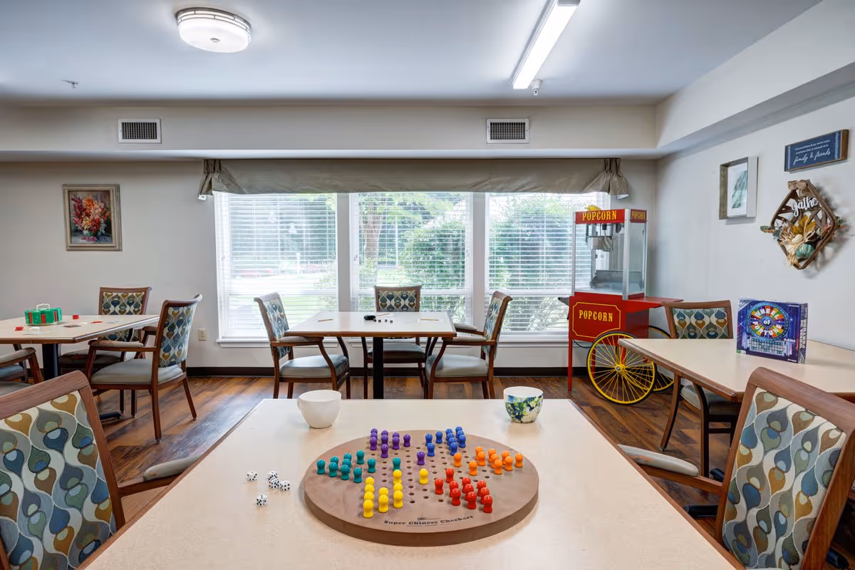 A bright activity room with several tables and chairs featuring patterned upholstery. On the nearest table, there is a colorful Chinese checkers game set up with dice and cups. In the background, there is a red popcorn machine and a board game on another table. Large windows with blinds allow natural light to fill the room, and the walls are decorated with framed pictures and a decorative wreath.