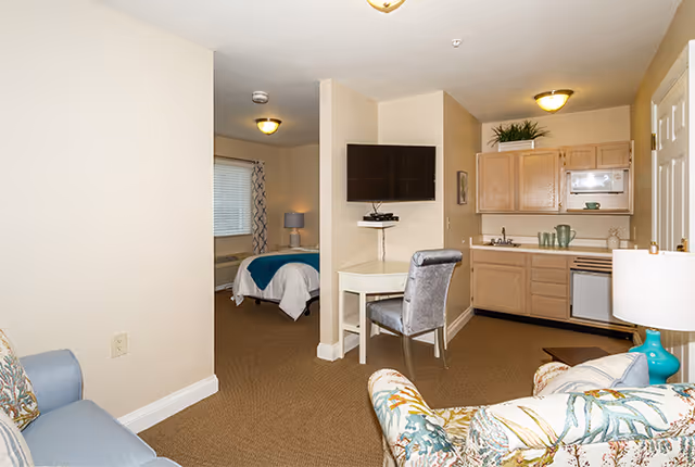 Interior view of a senior living facility unit showing a small living area with a floral armchair and a blue sofa, a wall-mounted TV above a small desk with a gray chair, a kitchenette with wooden cabinets, a microwave, and a mini fridge, and a bedroom with a bed covered in white and teal bedding visible through an open doorway.