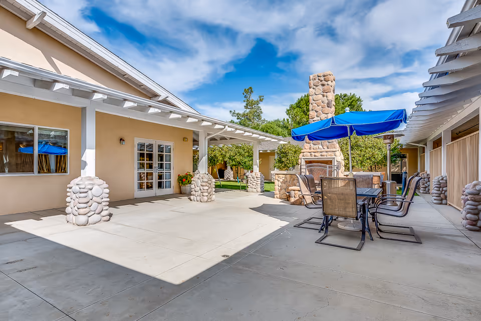 Sunny courtyard patio with an outdoor dining table and blue umbrella, stone fireplace, and covered walkways around it.
