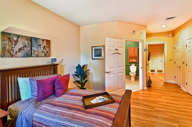 A cozy bedroom with a wooden bed frame, striped bedding, and multiple pillows in shades of purple and white. A wooden tray with items is placed on the bed. There is a plant next to the bed and a door open to a bathroom with a visible toilet. The room has wooden flooring and light-colored walls with framed artwork.