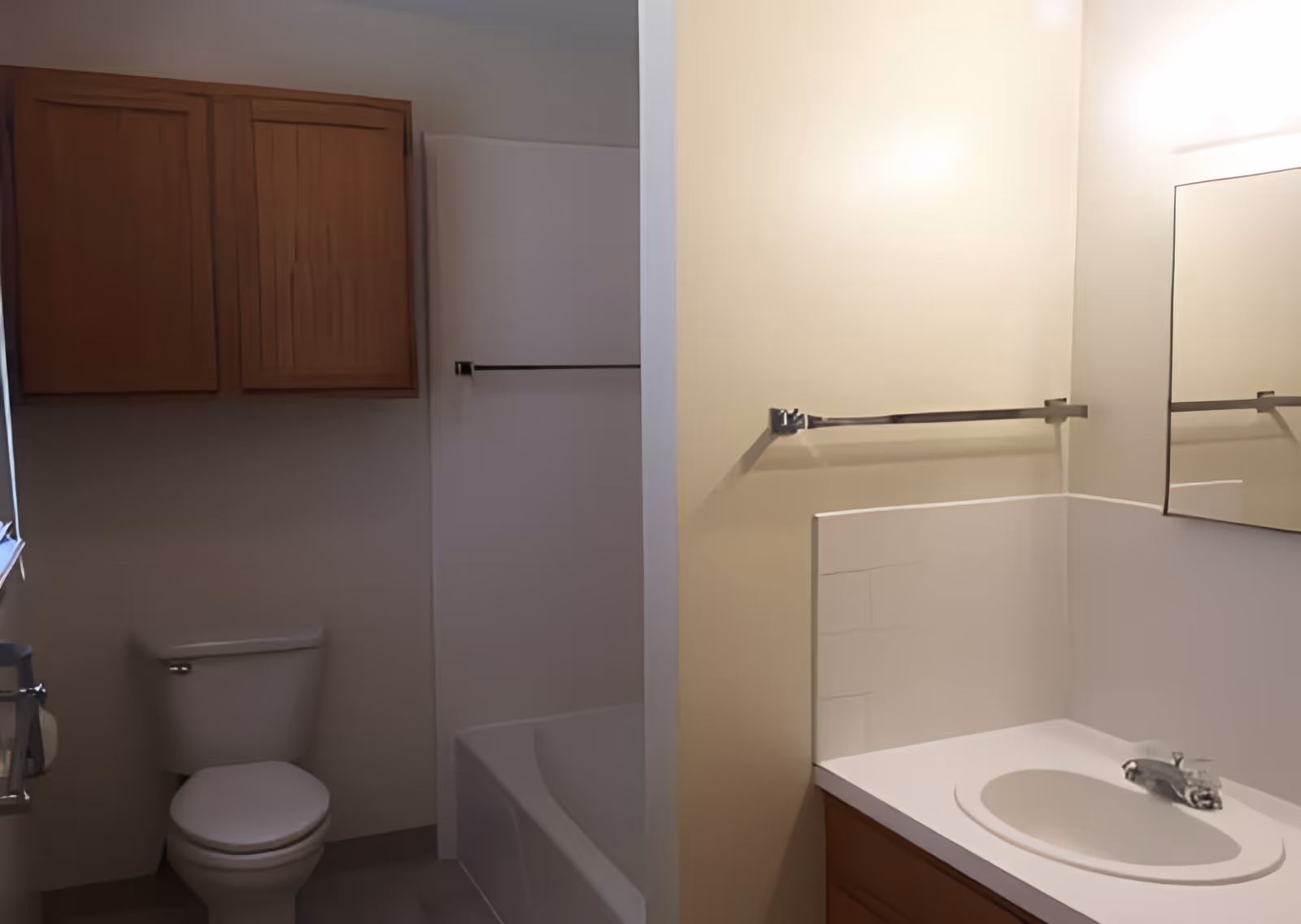 A small bathroom with a white toilet, a bathtub with a shower curtain rod, a wooden cabinet mounted on the wall above the toilet, a sink with a white countertop, a mirror above the sink, and a towel bar on the wall.