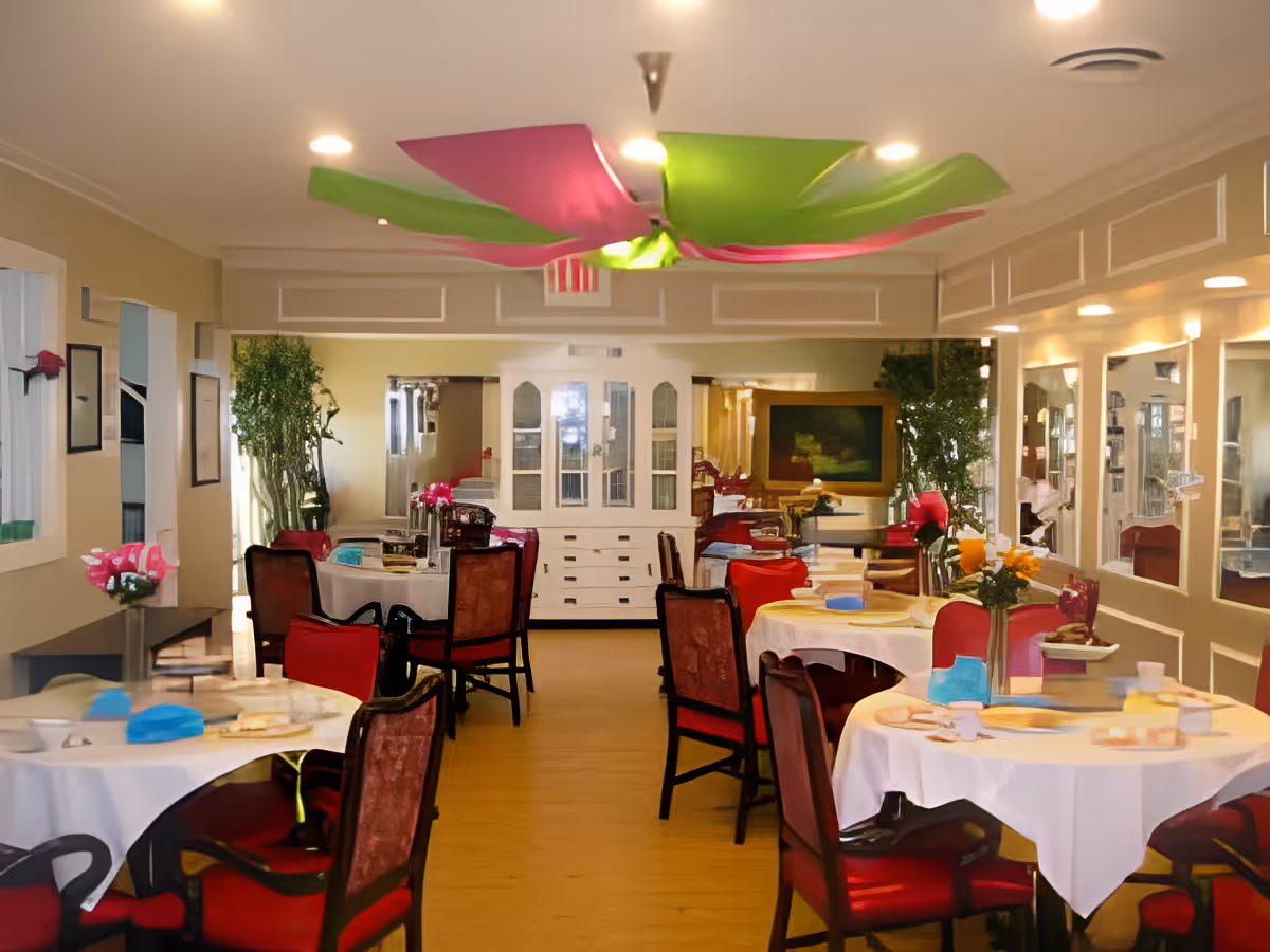 Dining room with round tables covered in white tablecloths, set with plates and napkins. Red cushioned chairs surround the tables. The room has beige walls with white trim, large mirrors on one side, and green plants near the back. Colorful pink and green fabric decorations hang from the ceiling.