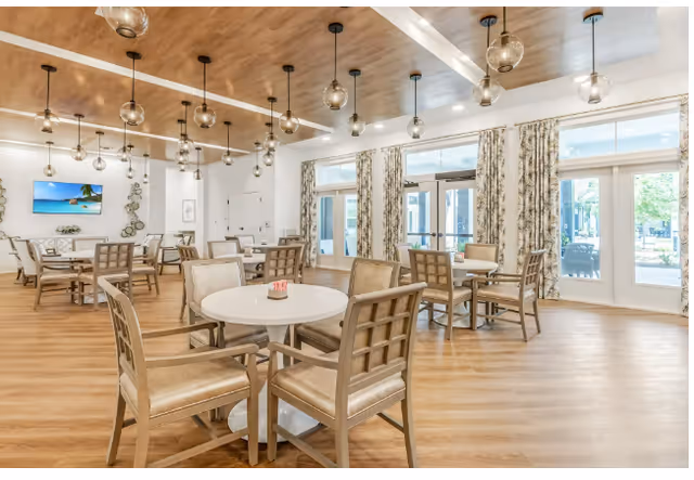 Spacious, well-lit communal dining room with round tables and wooden chairs, pendant lights, and large windows with patterned curtains.