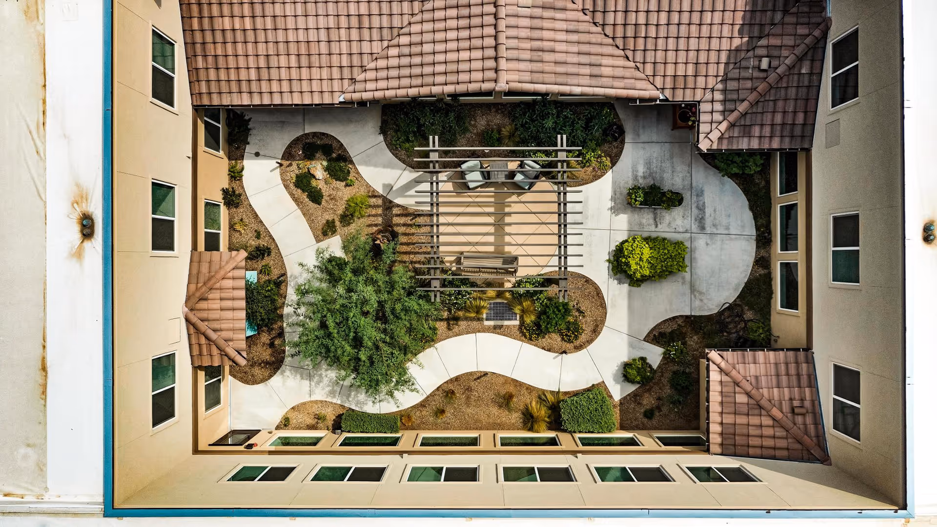 Aerial view of a courtyard in a senior living facility with a winding concrete pathway, landscaped garden beds, a pergola with seating underneath, and surrounding beige buildings with tiled roofs and multiple windows.