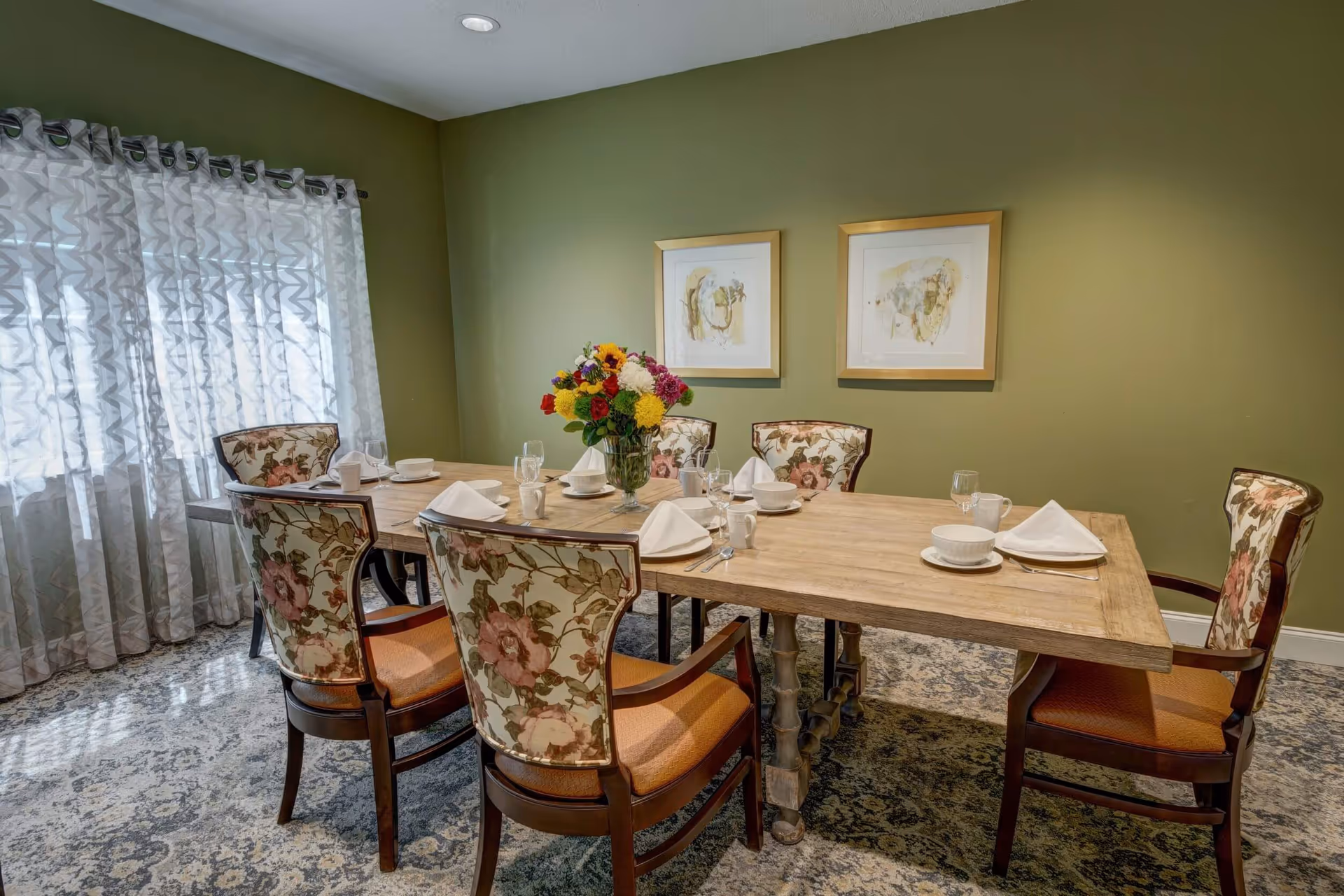 A dining room with a wooden table set for six people, featuring floral upholstered chairs, white napkins, cups, bowls, and glasses. A vase with colorful flowers is placed in the center of the table. The room has green walls with two framed artworks and a window with sheer patterned curtains allowing natural light in.