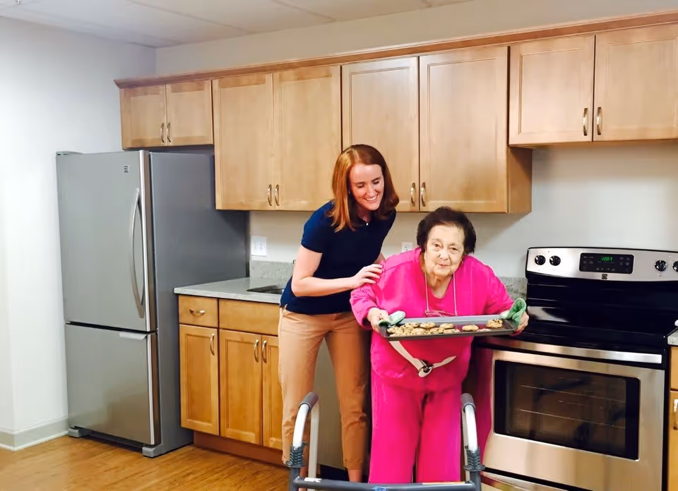 An elderly woman wearing a bright pink outfit is holding a tray of freshly baked cookies in a kitchen. A younger woman with red hair, dressed in a navy blue shirt and beige pants, is standing beside her, smiling and gently supporting her. The kitchen has wooden cabinets, a stainless steel refrigerator, and a stainless steel oven with a black cooktop.
