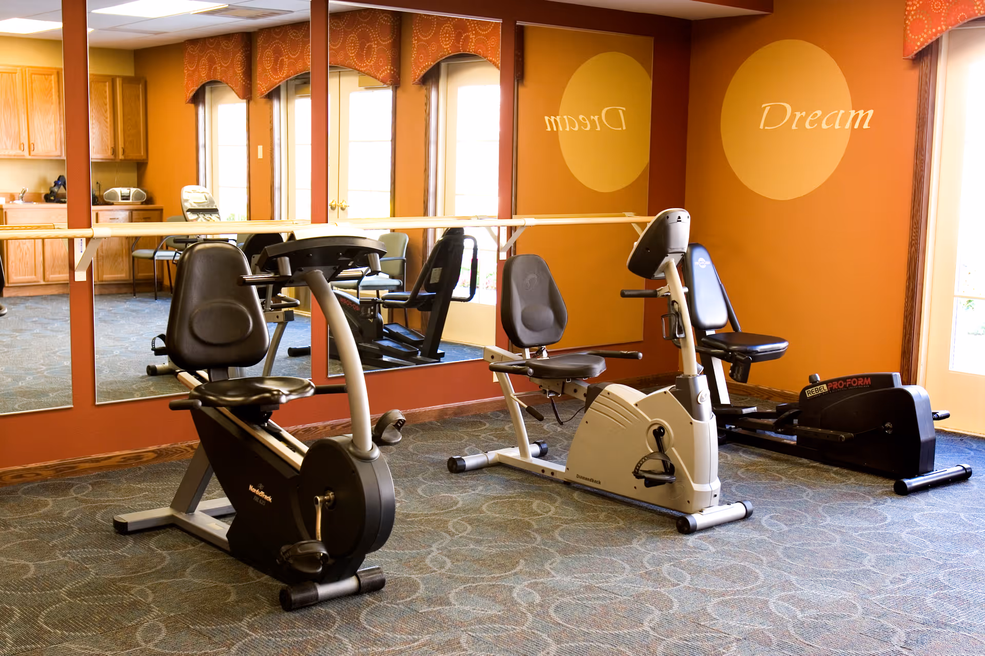 A small exercise room with three stationary exercise bikes positioned on a patterned carpet. The room has large mirrors on one wall reflecting the equipment and windows with orange valances. The word 'Dream' is painted on the orange wall near the windows.