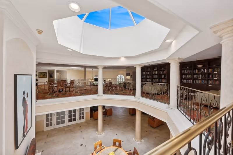 Two-level interior atrium with a central skylight, upper-level library and seating area overlooking a tiled ground floor with tables.