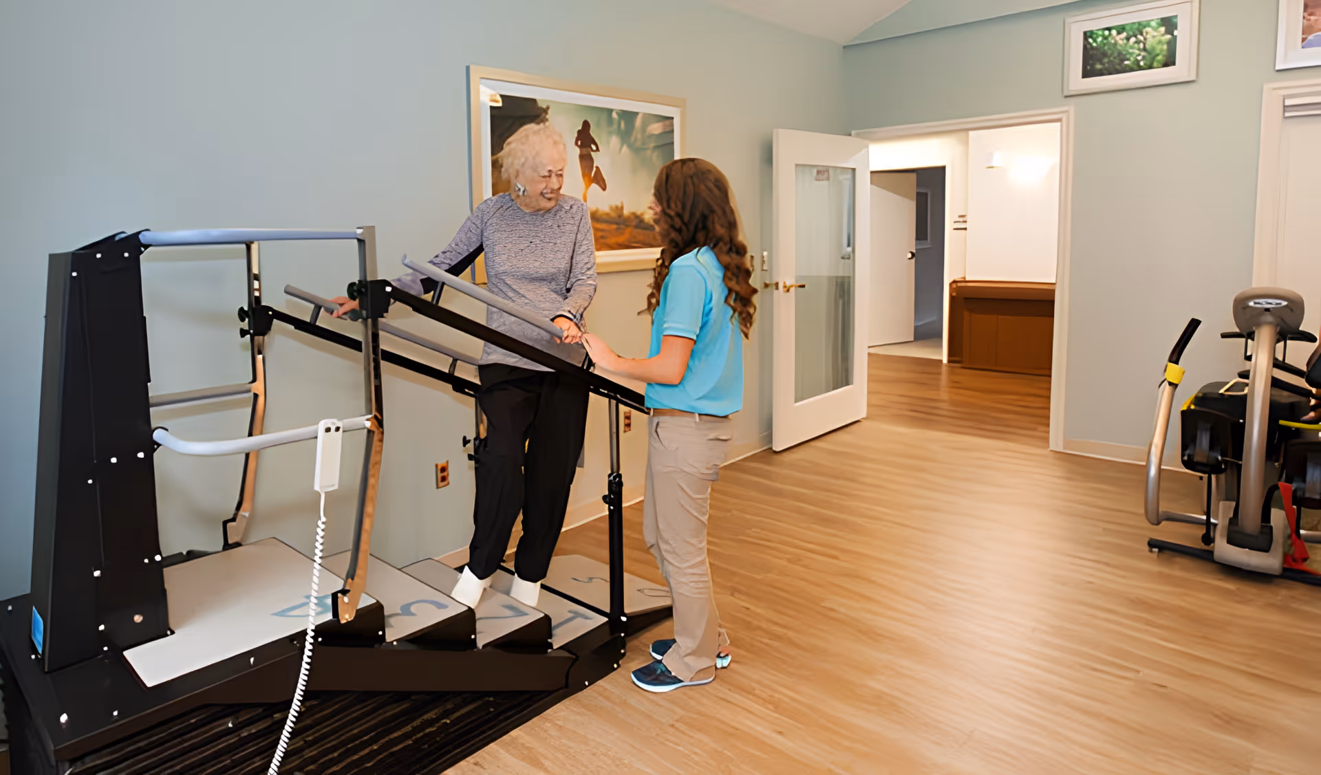 An elderly woman is assisted by a caregiver while using a stair exercise machine in a well-lit room with light blue walls and wooden flooring. Exercise equipment is visible in the background, and a framed picture hangs on the wall.