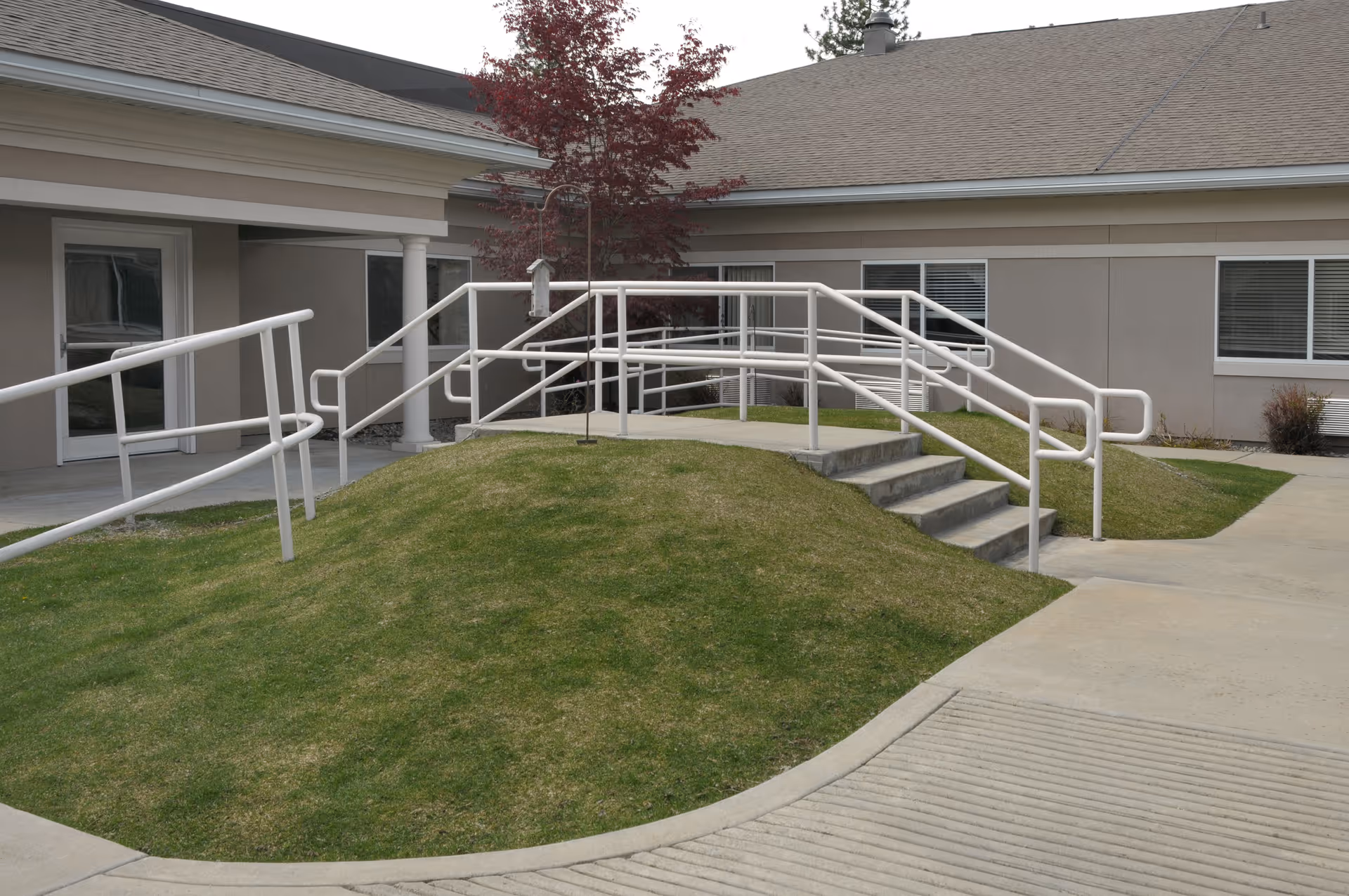 Outdoor area of a senior living facility featuring a small grassy hill with a concrete ramp and stairs equipped with white handrails, leading to a building entrance. The building has beige walls, white trim, and several windows with blinds. A small tree with red leaves is visible near the ramp.
