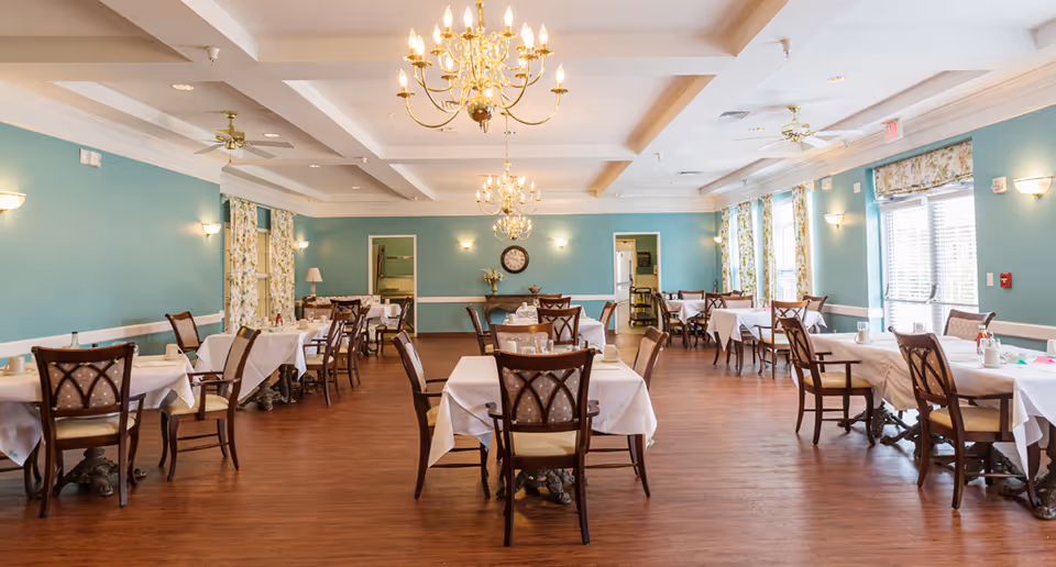 A spacious dining room with multiple tables covered in white tablecloths and wooden chairs arranged neatly. The walls are painted teal with floral curtains on the windows, and there are elegant chandeliers hanging from the ceiling. The floor is wooden, and the room is well-lit with natural light coming through the windows.