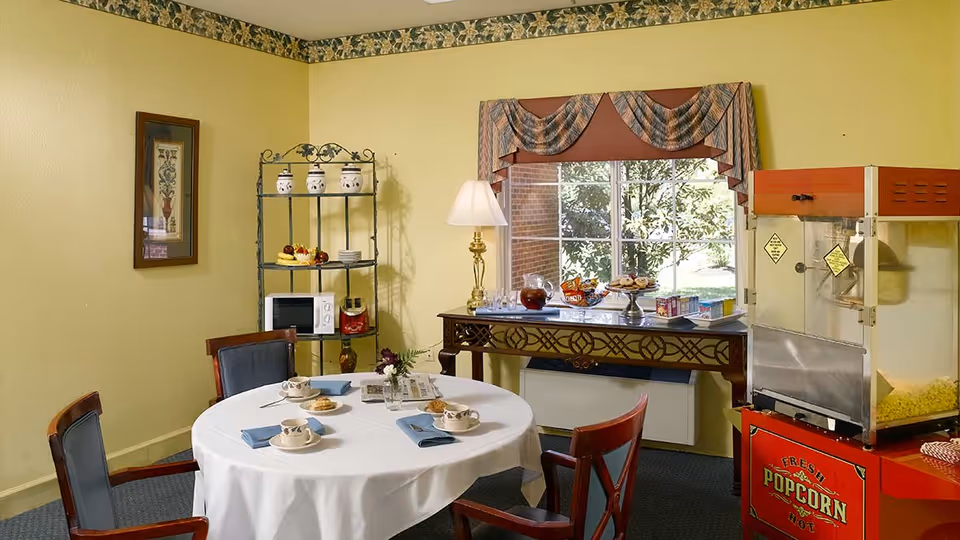 A small dining area with a round table covered with a white tablecloth, set with four cups and saucers, blue napkins, and cookies. Behind the table is a window with a decorative valance, a table with snacks and drinks, a popcorn machine, and a metal shelving unit holding a microwave and decorative jars. The walls are painted yellow with a floral border near the ceiling.