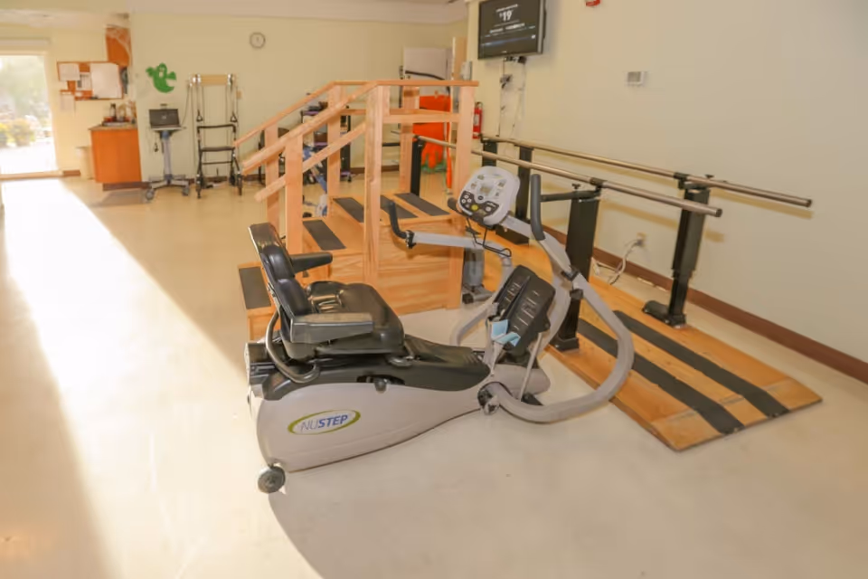 Physical therapy room with a NuStep recumbent cross trainer exercise machine, wooden stairs with handrails, and parallel bars for walking exercises in a rehabilitation center.