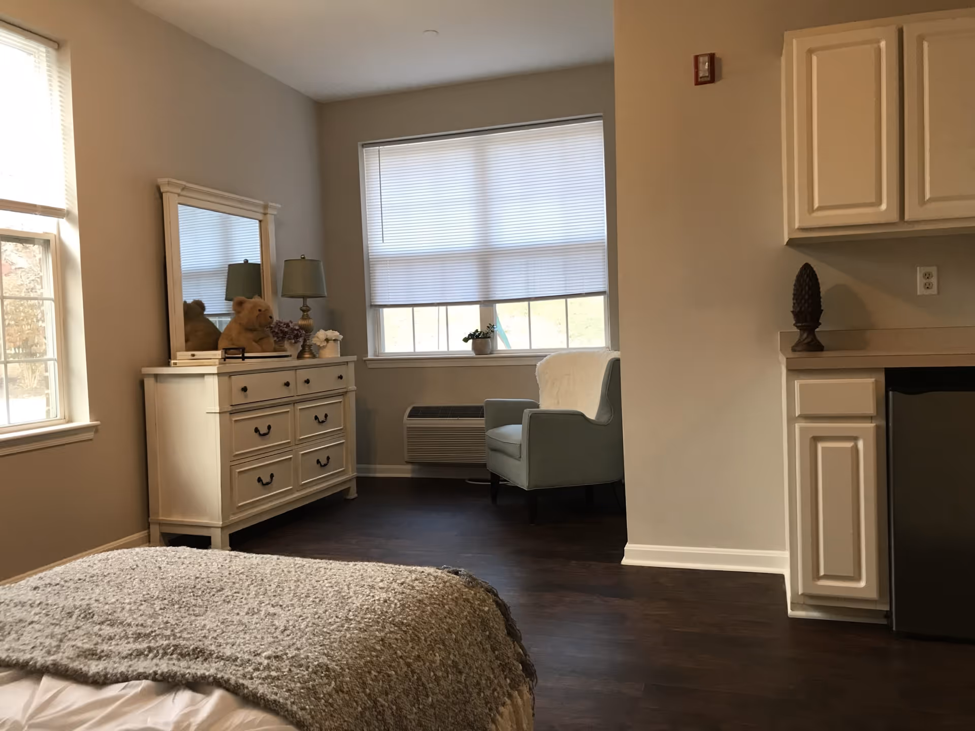 A cozy bedroom with a bed covered in a gray blanket in the foreground, a white dresser with a mirror and a teddy bear on top, a green armchair near a window with blinds, and white cabinets with a small refrigerator underneath on the right side.
