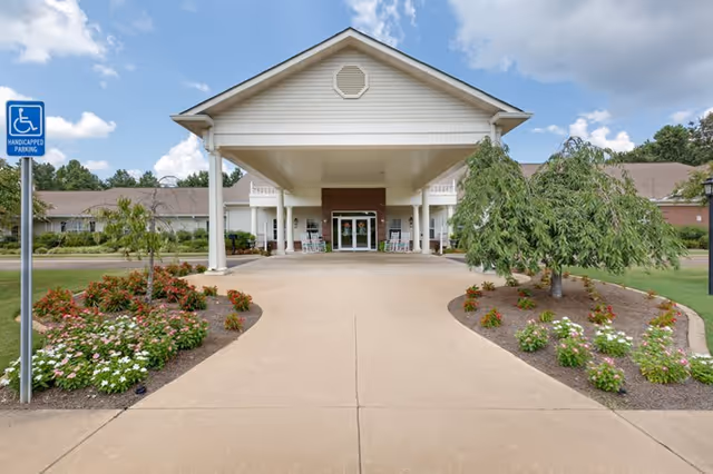 Front entrance of a single-story building with a covered driveway, landscaped flower beds on both sides, and a handicapped parking sign on the left. The building has white siding, a gabled roof, and a brick section around the entrance with glass doors and rocking chairs on the porch.