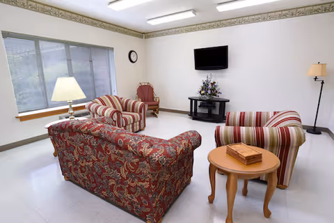 A bright living room with a large window on the left side. The room contains a patterned red sofa, a striped armchair, a matching striped loveseat, a wooden side table with a decorative box, a floor lamp, a table lamp on a small table near the window, a wall-mounted flat screen TV above a black TV stand with a floral arrangement, and a clock on the wall.