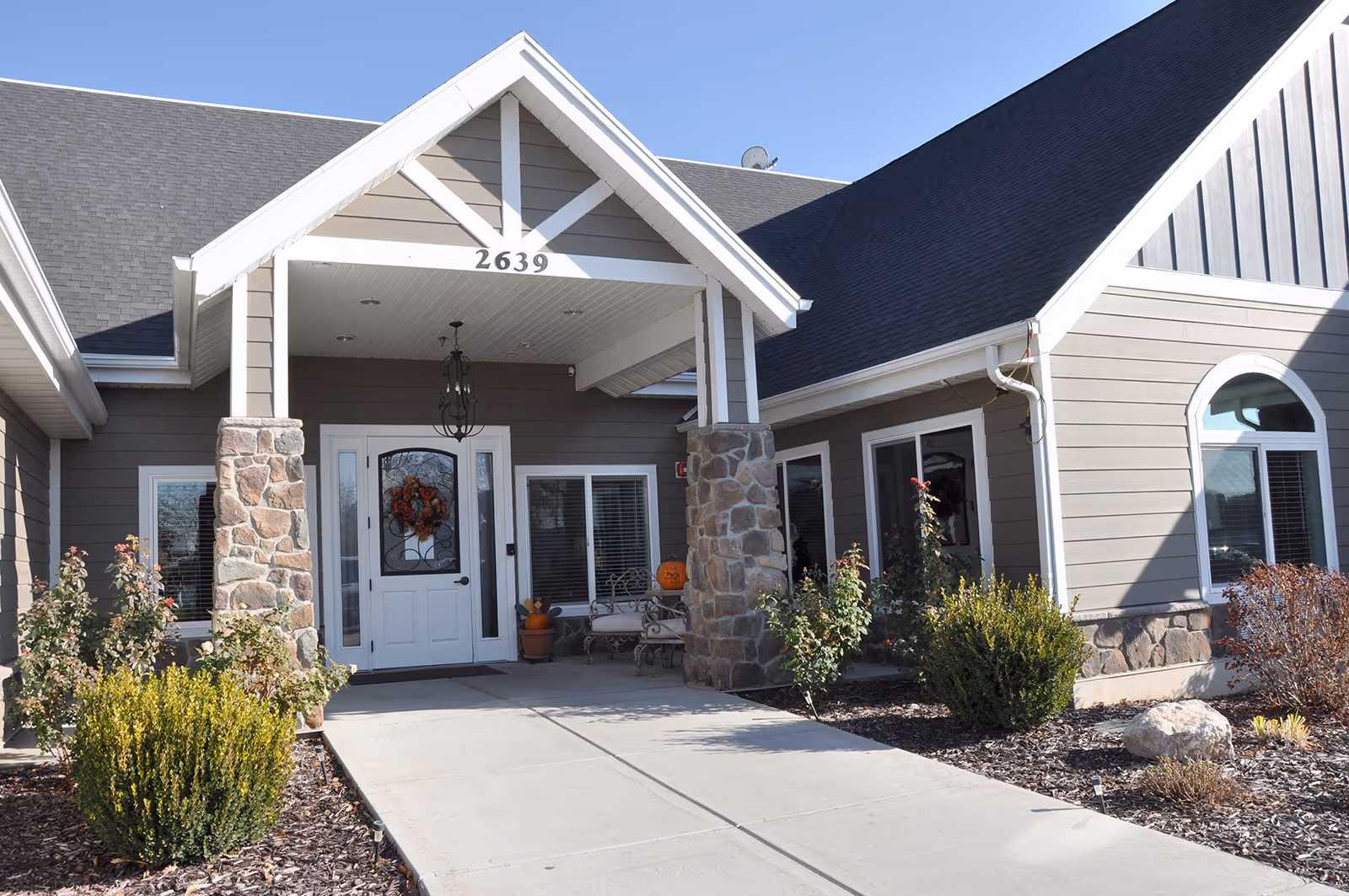 Exterior front entrance of a senior living facility with a covered porch supported by stone pillars, a white door decorated with a fall wreath, and surrounding landscaping with bushes and plants under a clear blue sky.