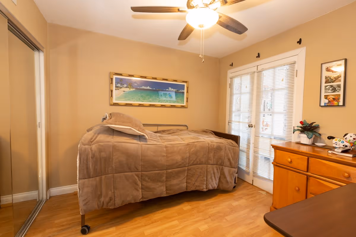 A cozy bedroom with a single bed covered in a brown blanket, a ceiling fan with light, a framed beach landscape picture on the wall, mirrored closet doors on the left, and a wooden dresser with a plant and decorative items on the right. French doors with blinds are visible in the background.
