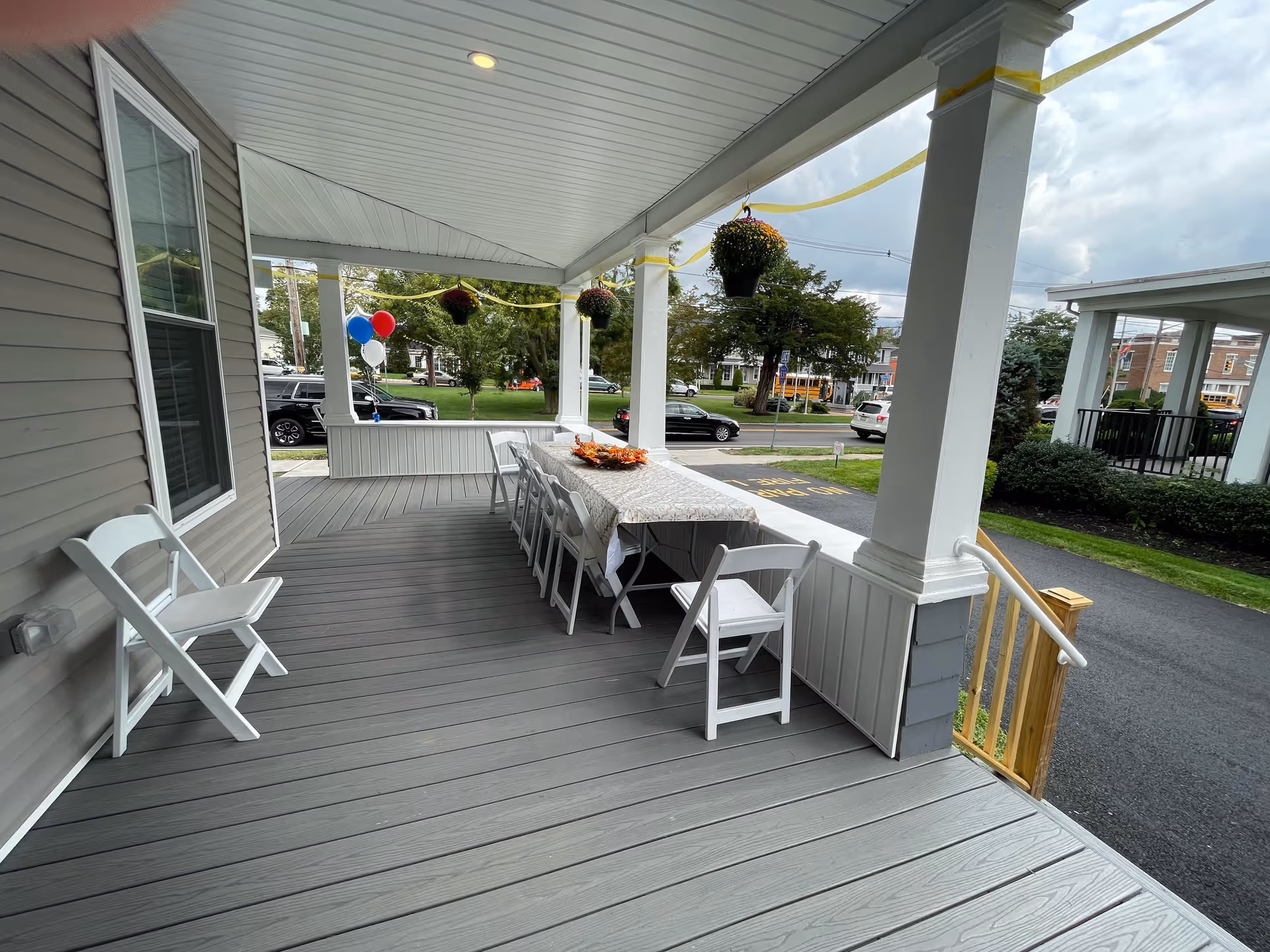 A covered outdoor porch area with gray wooden flooring and white pillars. There is a long table covered with a patterned tablecloth and decorated with a centerpiece, surrounded by white folding chairs. Hanging flower pots and balloons in red, white, and blue are also visible. The porch overlooks a street with parked cars and trees.