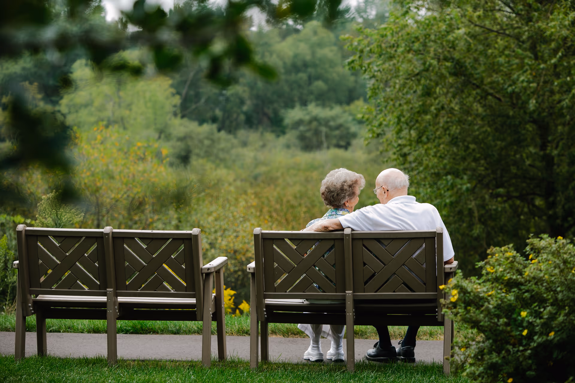 An elderly couple sitting closely together on a wooden bench in a peaceful outdoor garden area surrounded by green trees and bushes.