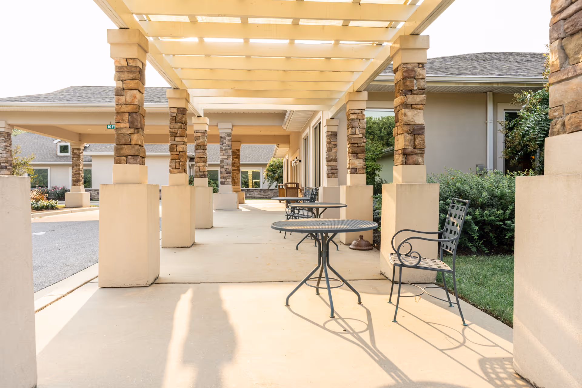 Covered outdoor seating area with stone pillars, metal tables and chairs along a walkway at a senior living facility.