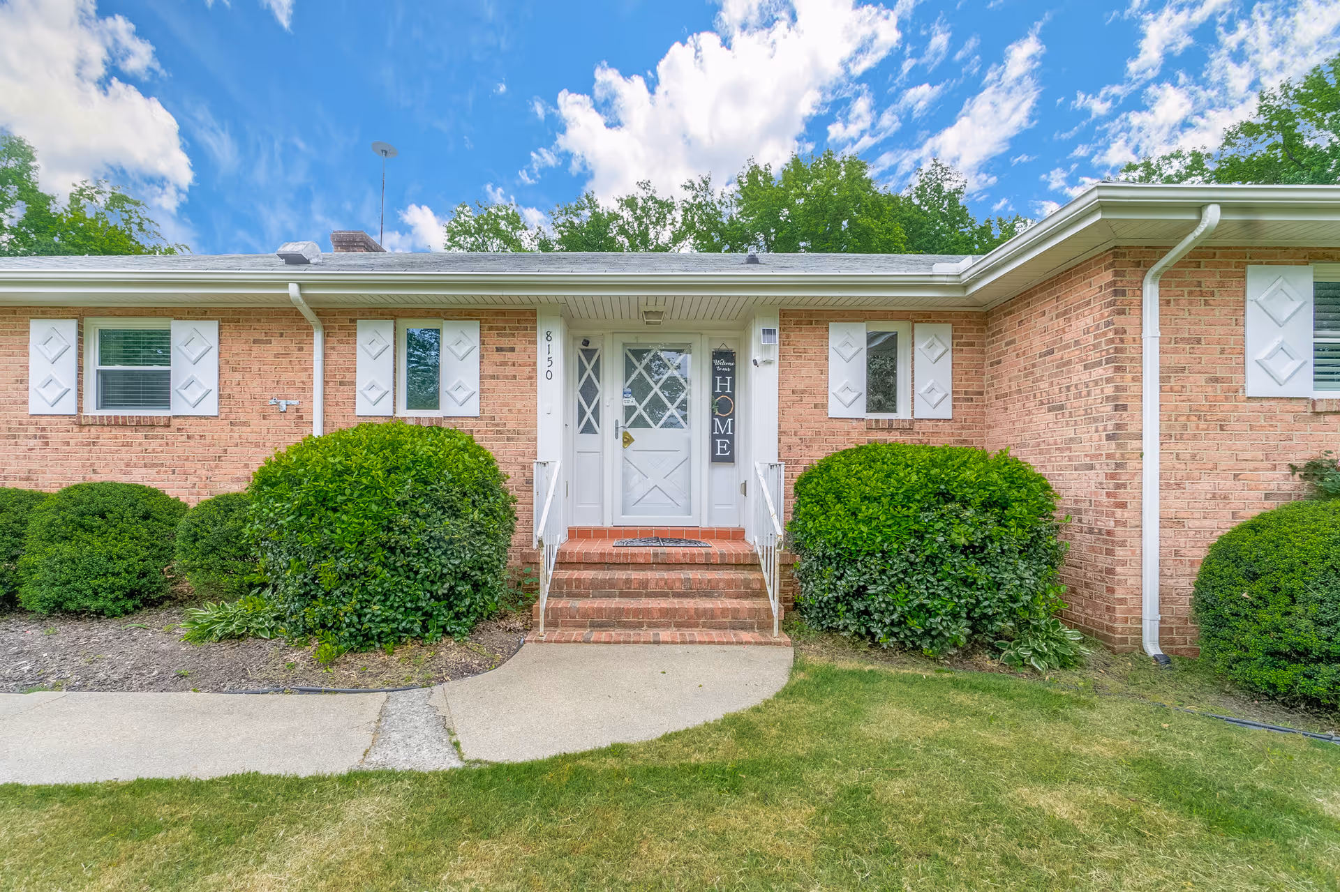 Front exterior view of a single-story brick building with white shutters and a white front door. There are green bushes on either side of the entrance and a concrete walkway leading up to the door. The sky is blue with some clouds.