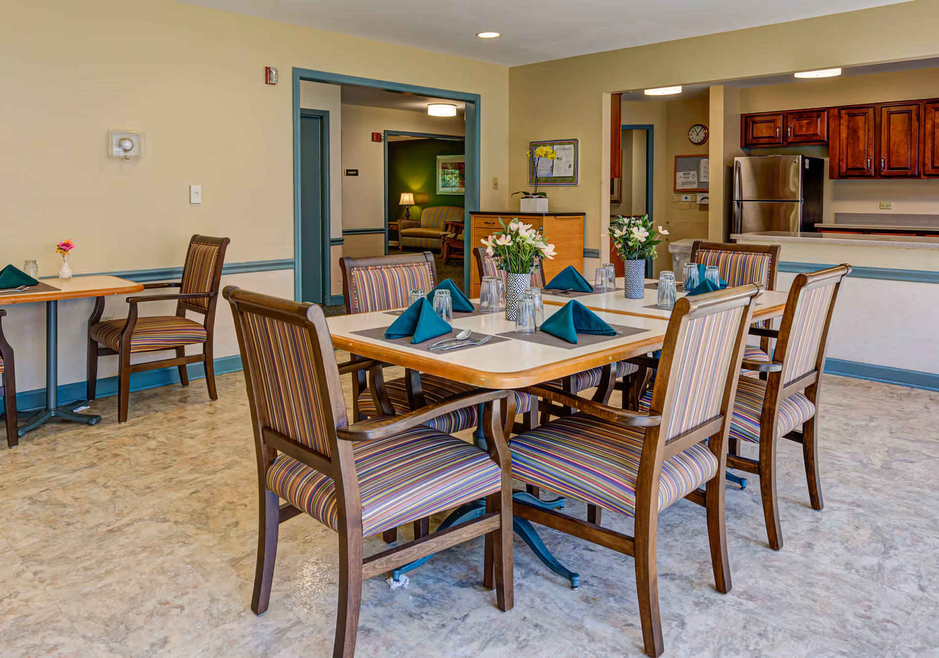 A dining area in a senior living facility with a rectangular table set for six people. The table has striped wooden chairs, green folded napkins, glasses, and vases with flowers. In the background, there is a kitchen area with wooden cabinets and a stainless steel refrigerator, and an adjoining room with a sofa and lamp is visible through a doorway.