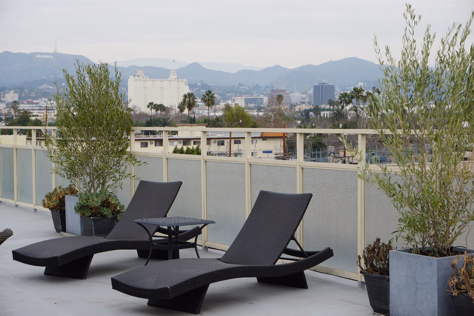 Outdoor patio area with two black woven lounge chairs and a small matching table between them. There are potted plants on either side of the chairs. In the background, a cityscape with buildings, palm trees, and distant mountains is visible under a cloudy sky.