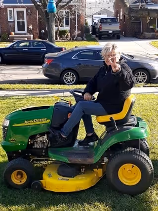 A person sitting on a green and yellow John Deere riding lawn mower on a grassy lawn in a residential neighborhood. The person is waving and smiling. There are cars parked on the street and houses in the background.