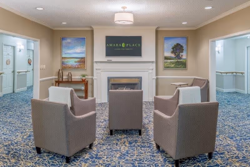 A cozy seating area in a senior living facility with four gray armchairs arranged around a fireplace. Above the fireplace is a TV screen displaying the text 'AMARA PLACE'. The room has beige walls, two landscape paintings on either side of the fireplace, and a blue patterned carpet. There are hallways visible on both sides of the seating area.