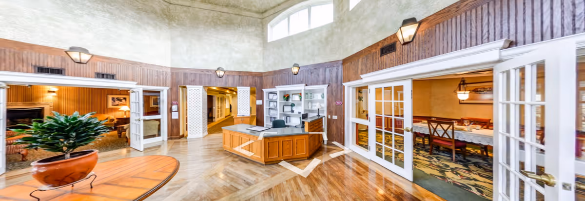 Interior view of a senior living facility lobby area with a wooden reception desk in the center, surrounded by wood-paneled walls and multiple doorways leading to other rooms. To the left, there is a cozy lounge area with a fireplace and seating, and to the right, a dining room with tables and chairs visible through glass-paned double doors. The floor is wooden with a patterned carpet in the dining area, and the ceiling is high with wall-mounted lights.
