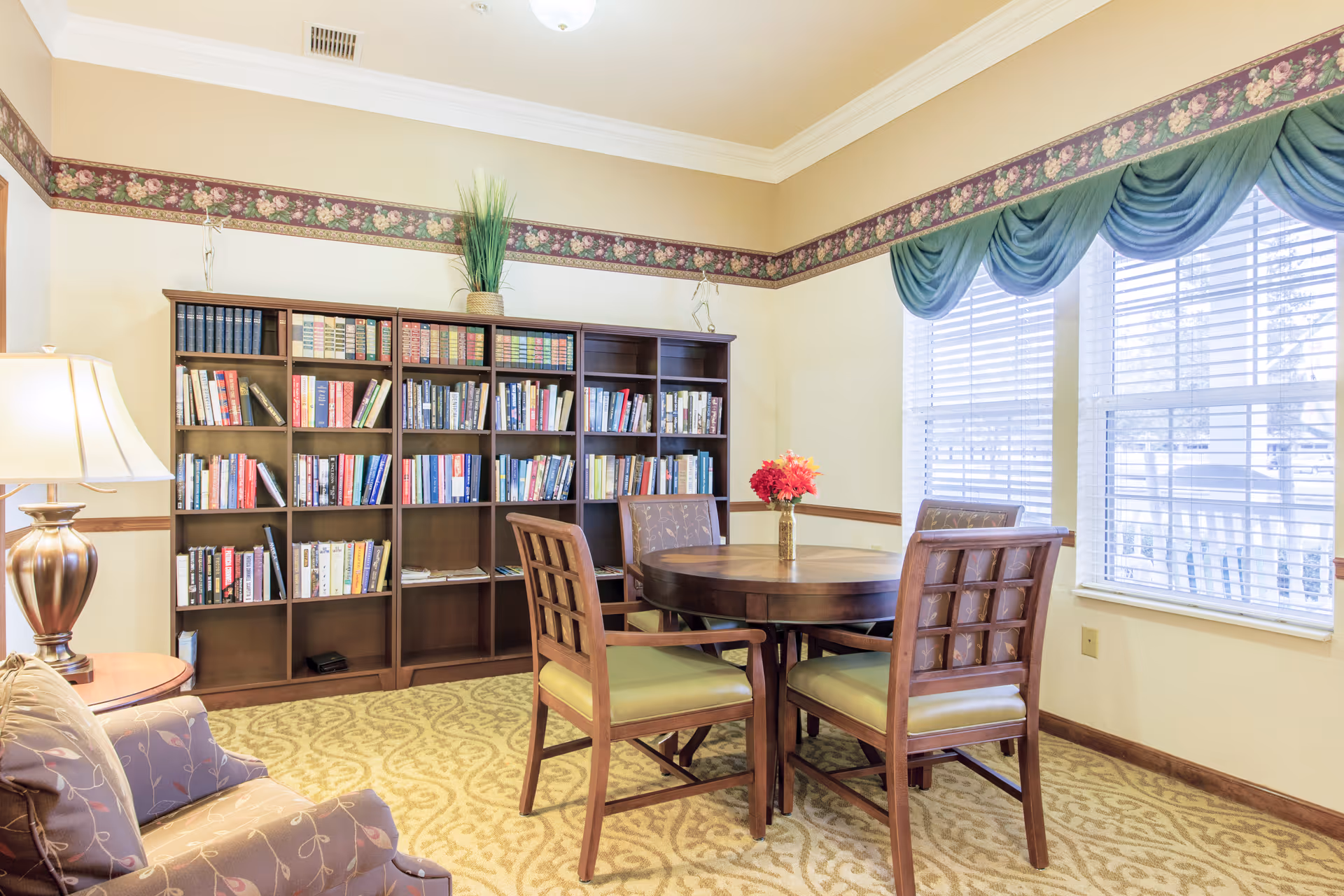 A cozy interior room with a round wooden table surrounded by four wooden chairs with green cushions. Behind the table is a large bookshelf filled with books and a decorative plant on top. To the left, there is a cushioned armchair with a patterned fabric and a table lamp on a small side table. The room has beige walls with a floral wallpaper border near the ceiling and large windows with green drapes letting in natural light.
